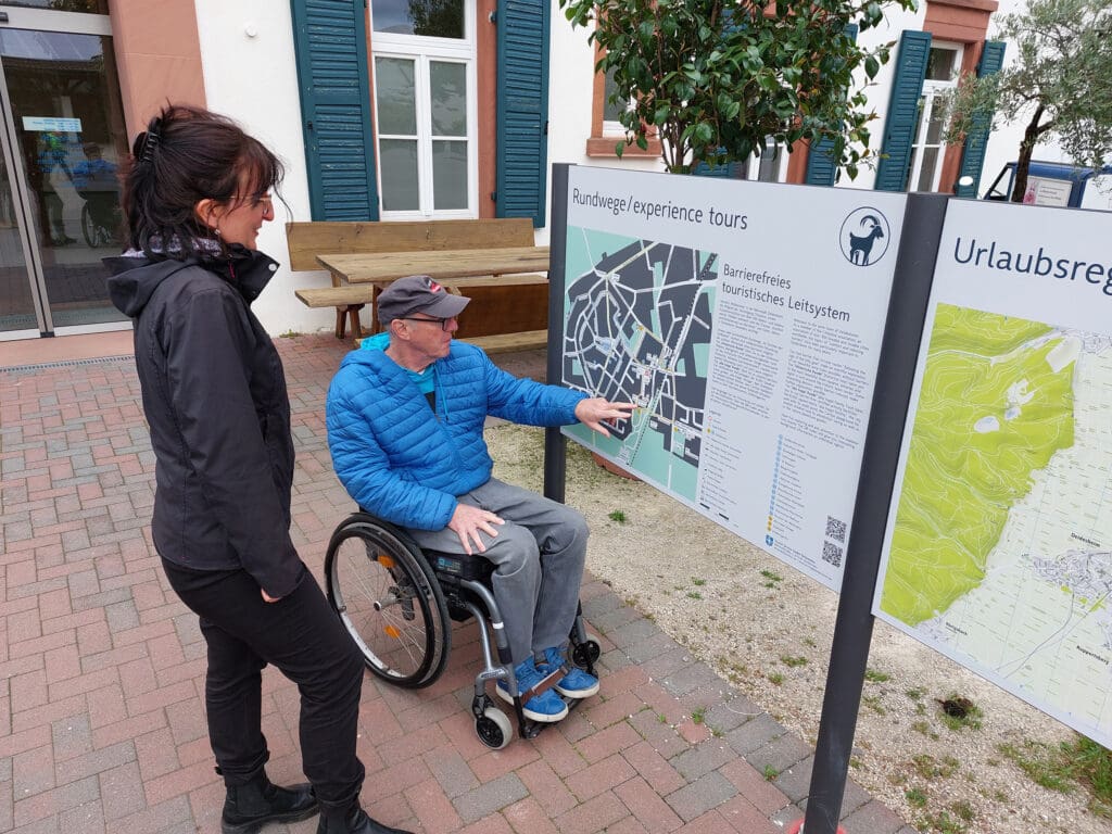 Ein Mann im Rollstuhl schaut sich das barrierefreie touristische Leitsystem auf einer Infotafel vor dem Deidesheimer Rathaus an